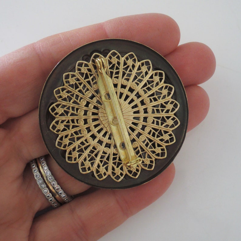 Peut inclure: Une broche ronde noire et dor&eacute;e avec un motif d&eacute;coratif en filigrane. La broche a un fermoir &agrave; &eacute;pingle.