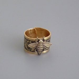 May include: A gold-toned ring with a detailed bee design and floral accents. The ring is made of metal and has a wide band.