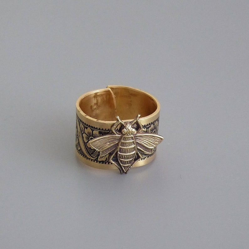 May include: A gold-toned ring with a detailed bee design and floral accents. The ring is made of metal and has a wide band.