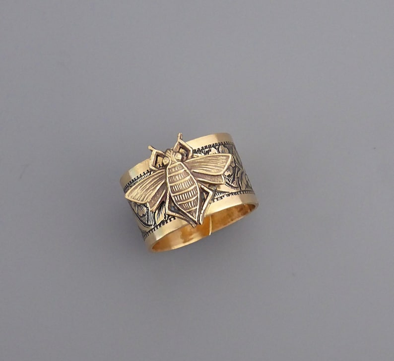May include: A gold ring with a detailed bee design. The bee is raised and has a textured body. The ring band is engraved with a floral pattern.