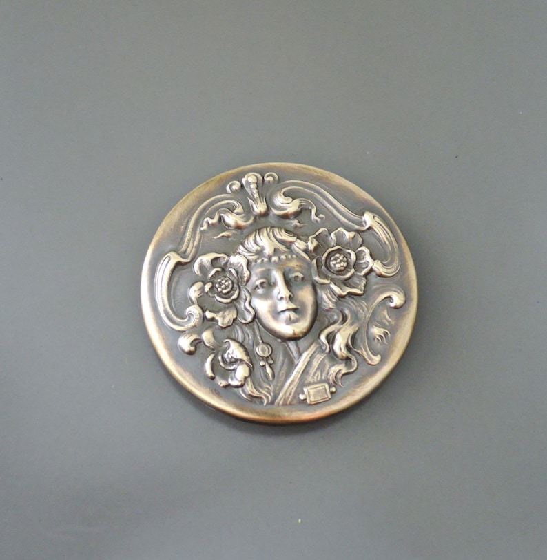 Peut inclure: Une broche ronde argent&eacute;e avec un motif en relief d'un visage de femme entour&eacute; de motifs floraux.
