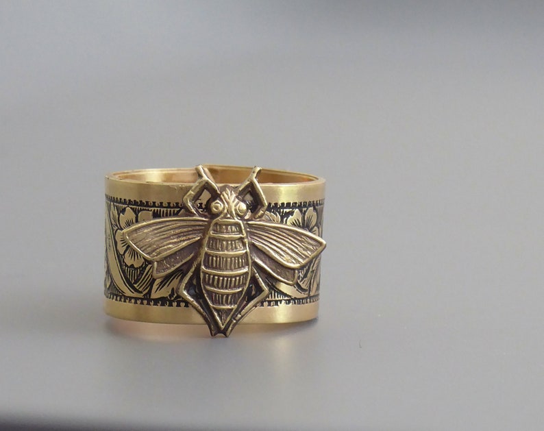 May include: A gold-toned metal ring with a detailed bee design. The ring is decorated with an intricate floral pattern.