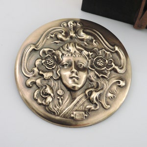 Peut inclure: Une broche ronde en laiton avec un motif en relief d'un visage de femme entour&eacute; de motifs floraux.