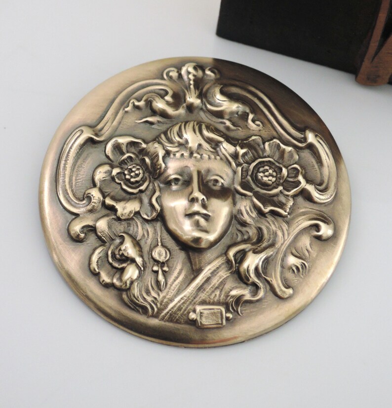 Peut inclure: Une broche ronde en laiton avec un motif en relief d'un visage de femme entour&eacute; de motifs floraux.
