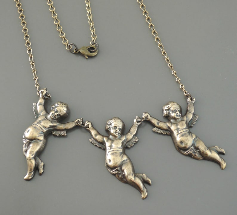 May include: A gold chain necklace with three cherub figures holding hands. The cherubs are in a flying position and have wings.