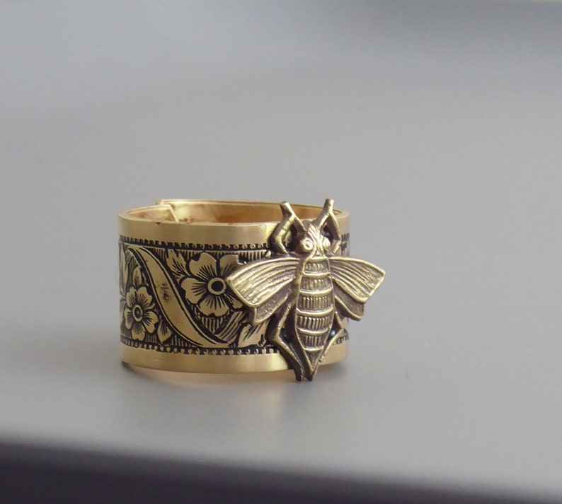 May include: A gold-toned ring with a detailed floral design and a bee charm. The bee is three-dimensional and has a textured surface.