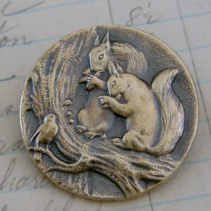 May include: A round, antique-style button with a detailed relief design. The button features two squirrels interacting on a tree branch, with a small bird perched nearby. The button has a bronze-like finish and is set against a textured background.