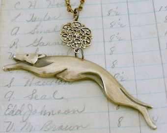 May include: A gold necklace with a detailed filigree pendant and a stylized greyhound pendant. The necklace is lying on a piece of lined paper with handwritten text.