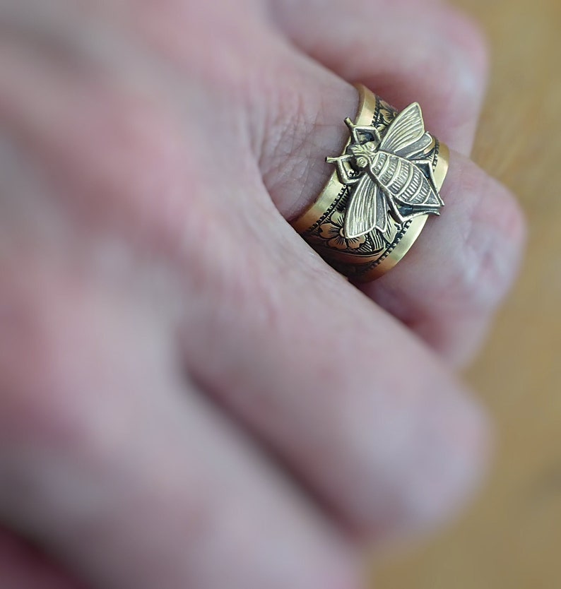 May include: A gold-toned ring with a detailed bee design. The ring is worn on a finger and has a floral pattern on the band.