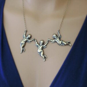 May include: A gold-toned necklace with three cherub figures holding hands. The cherubs are in a flying pose and are attached to a delicate chain.