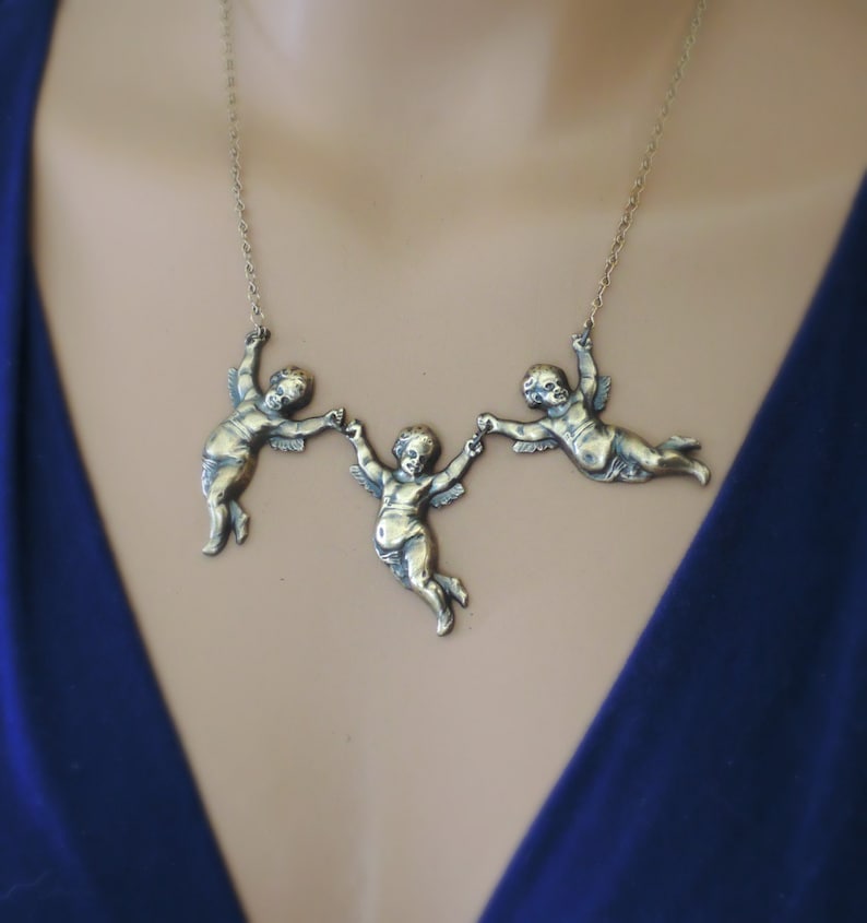 May include: A gold-toned necklace with three cherub figures holding hands. The cherubs are in a flying pose and are attached to a delicate chain.