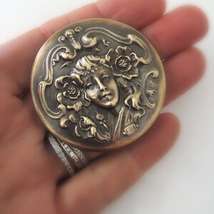 Peut inclure: Un compact rond en laiton avec un design orn&eacute; repr&eacute;sentant un visage de femme entour&eacute; de fleurs et de volutes. Le compact a un aspect l&eacute;g&egrave;rement vieilli, antique.
