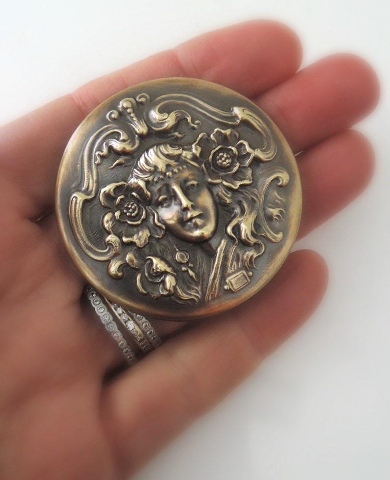 Peut inclure: Un compact rond en laiton avec un design orn&eacute; repr&eacute;sentant un visage de femme entour&eacute; de fleurs et de volutes. Le compact a un aspect l&eacute;g&egrave;rement vieilli, antique.