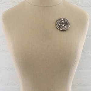 Peut inclure: Une broche ronde en bronze avec un motif en relief d'un visage de femme entour&eacute; de motifs floraux.