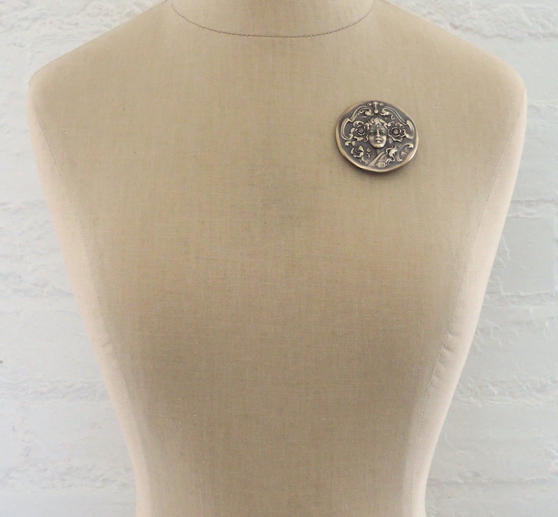 Peut inclure: Une broche ronde en bronze avec un motif en relief d'un visage de femme entour&eacute; de motifs floraux.