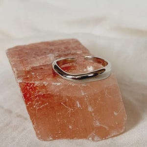 Könnte beinhalten: Ein silberner Ring mit einer einzigartigen, unregelmäßigen Form liegt auf einem rechteckigen, hellorangen Stein. Die polierte Oberfläche des Rings reflektiert das Licht und kontrastiert mit der strukturierten Oberfläche des Steins. Der Hintergrund ist ein weicher, weißer Stoff.