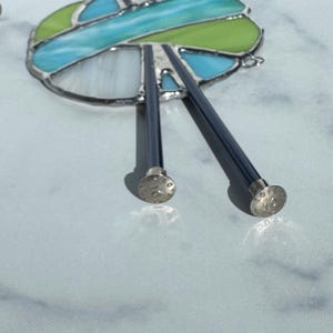 May include: A pair of dark blue knitting needles with silver-colored tips, resting on a light blue surface. The needles are positioned near a stained glass piece with green, blue, and white sections, outlined in silver.