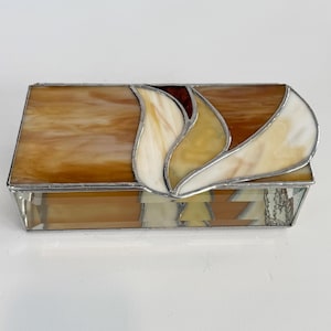 May include: A rectangular stained glass box with a lid featuring a stylized floral design. The box has a warm color palette of amber, cream, and brown, with a silver metal frame. The design includes a layered, curved shape on the lid.
