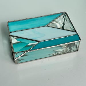 May include: A rectangular stained glass box with a geometric design. The box features turquoise, light blue, and clear glass panels, all framed with a silver-colored metal. The box is likely used for storing small items.
