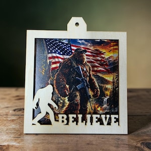 May include: Wooden framed art piece featuring a Bigfoot-like creature holding a rifle in front of an American flag. The word "BELIEVE" is cut out at the bottom. A Bigfoot silhouette is on the left side. The background shows a mountain landscape.