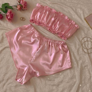 May include: A light pink satin pajama set, including a ruffled tube top and matching shorts, is displayed on a bed. The fabric has a shiny, smooth texture. Pink roses and gold jewelry are arranged nearby.