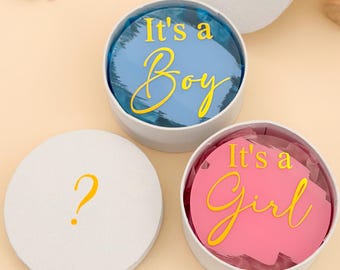 Könnte beinhalten: Weiße runde Schachteln mit goldenen Fragezeichen und Botschaften zur Geschlechtsenthüllung. Eine Schachtel zeigt "It's a Boy" auf blauem Hintergrund, eine andere "It's a Girl" auf rosa. Eine dritte Schachtel hat ein Fragezeichen.