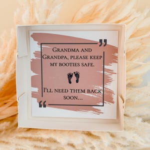 Puede incluir: Una caja de regalo cuadrada blanca que contiene una tarjeta con un fondo de pincelada rosa polvoriento. El texto dice: Grandma and Grandpa, please keep my booties safe. I'll need them back soon... con dos peque&ntilde;as huellas de beb&eacute; negras.