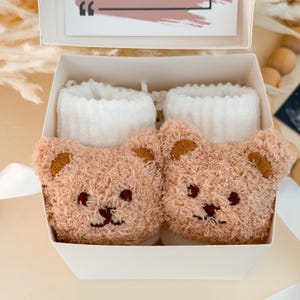 Puede incluir: Un par de calcetines beige difusos con un dise&ntilde;o de cara de oso de peluche en la parte delantera, con ojos y nariz bordados en marr&oacute;n oscuro, junto con pu&ntilde;os acanalados blancos, presentados dentro de una caja de regalo blanca con una tarjeta.