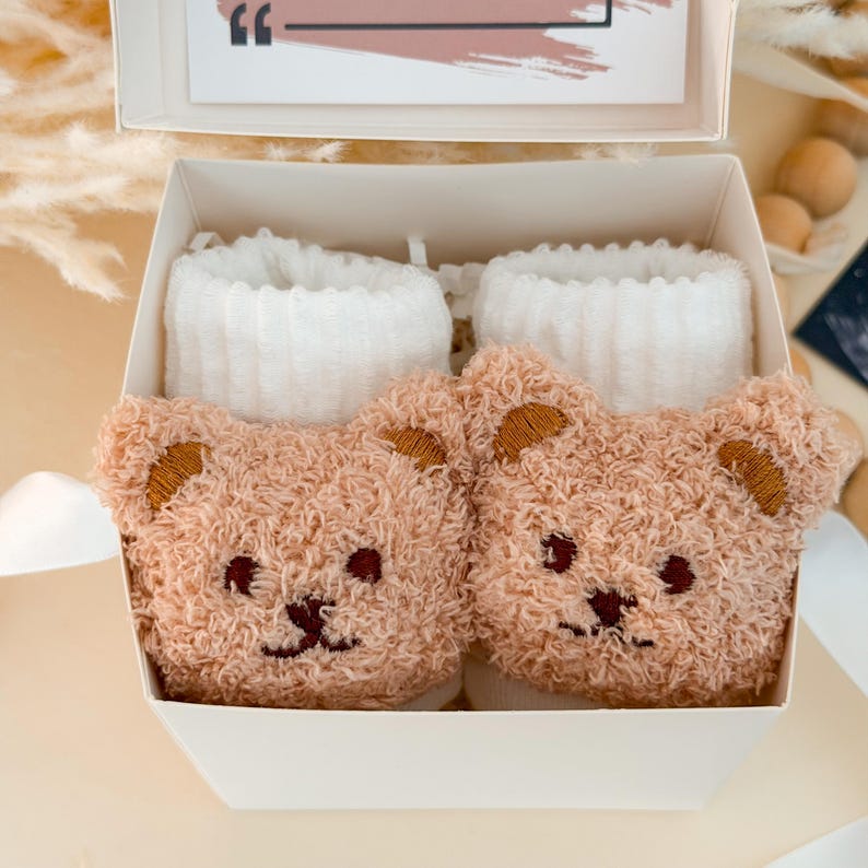 Puede incluir: Un par de calcetines beige difusos con un dise&ntilde;o de cara de oso de peluche en la parte delantera, con ojos y nariz bordados en marr&oacute;n oscuro, junto con pu&ntilde;os acanalados blancos, presentados dentro de una caja de regalo blanca con una tarjeta.