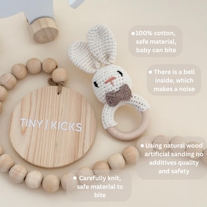 Puede incluir: Un sonajero de conejo tejido a ganchillo con un anillo de madera, que presenta una cabeza de color crema, orejas largas y una pajarita marr&oacute;n. Junto a &eacute;l hay una placa de madera circular con el texto TINY | KICKS y un collar de cuentas de madera natural.