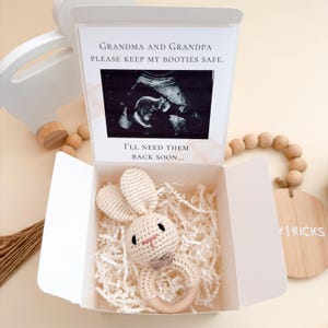 May include: A white box with a crochet bunny rattle and a sonogram image. The box contains shredded paper and a crochet bunny rattle with a wooden ring. The text on the box reads: "Grandma and Grandpa, Please keep my booties safe. I'll need them back soon..."
