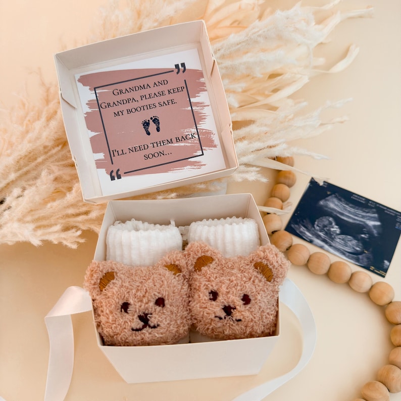 Puede incluir: Una caja de regalo blanca que contiene dos patucos de beb&eacute; de peluche color canela con forma de oso y pu&ntilde;os acanalados blancos. La tapa incluye una tarjeta con el texto: Grandma and Grandpa, please keep my booties safe. I'll need them back soon.