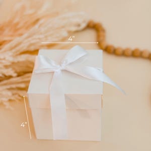 Puede incluir: Una caja de regalo cuadrada de color blanco con un lazo de sat&eacute;n blanco atado en la parte superior. La caja muestra unas dimensiones de 10,16 cm de ancho y 10,16 cm de alto, indicadas claramente con l&iacute;neas y texto blanco en la imagen.