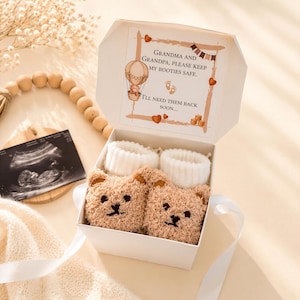 Puede incluir: Una caja de regalo blanca que contiene dos patucos de beb&eacute; de color beige con forma de oso y pu&ntilde;os blancos acanalados. La tapa de la caja incluye el mensaje impreso: Grandma and Grandpa, please keep my booties safe. I'll need them back soon.