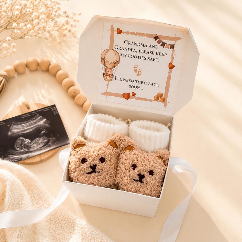 Puede incluir: Una caja de regalo blanca que contiene dos patucos de beb&eacute; de color beige con forma de oso y pu&ntilde;os blancos acanalados. La tapa de la caja incluye el mensaje impreso: Grandma and Grandpa, please keep my booties safe. I'll need them back soon.