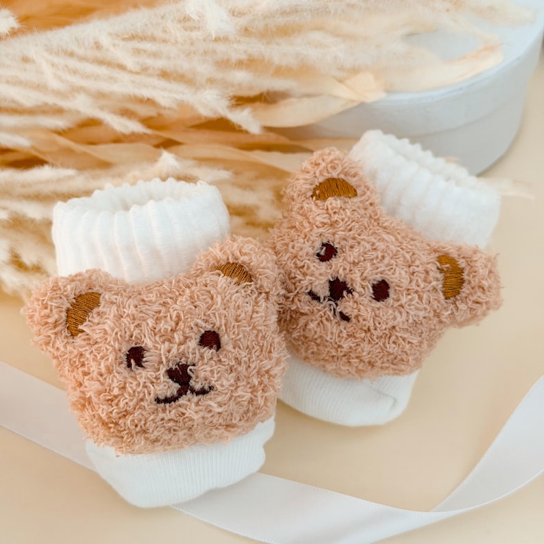 Puede incluir: Un par de calcetines blancos con una cara de oso de peluche de color marr&oacute;n claro texturizado en la parte delantera. El dise&ntilde;o del oso incluye ojos, nariz y boca bordados en marr&oacute;n oscuro, con peque&ntilde;as orejas redondeadas en los laterales.