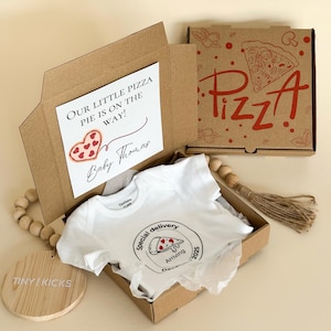 Puede incluir: Un juego de regalo para bebé en una caja de cartón, que incluye un body blanco con un gráfico de pizza y el texto "Special delivery Arriving Dec 2025". La caja también contiene una tarjeta que dice "Our little pizza pie is on the way!" y "Baby Thomas".