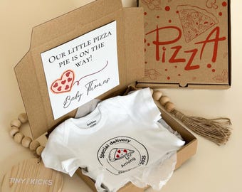 May include: A baby gift set in a cardboard box, including a white onesie with a pizza graphic and the text "Special delivery Arriving Dec 2025". The box also contains a card that says "Our little pizza pie is on the way!" and "Baby Thomas".