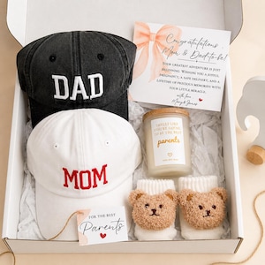 May include: A gift box containing a black baseball cap with white DAD embroidery, a white baseball cap with red MOM embroidery, a candle labeled Smells Like You're Going To Be The Best Parents, two white baby socks with brown bear faces, and cards.