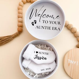 May include: A white gift box with the text "Welcome to your Auntie Era" and baby footprints. Inside, white baby socks with "I love Auntie Jessica" and "Est. 2026" are nestled in paper filler. A wooden disc is visible.