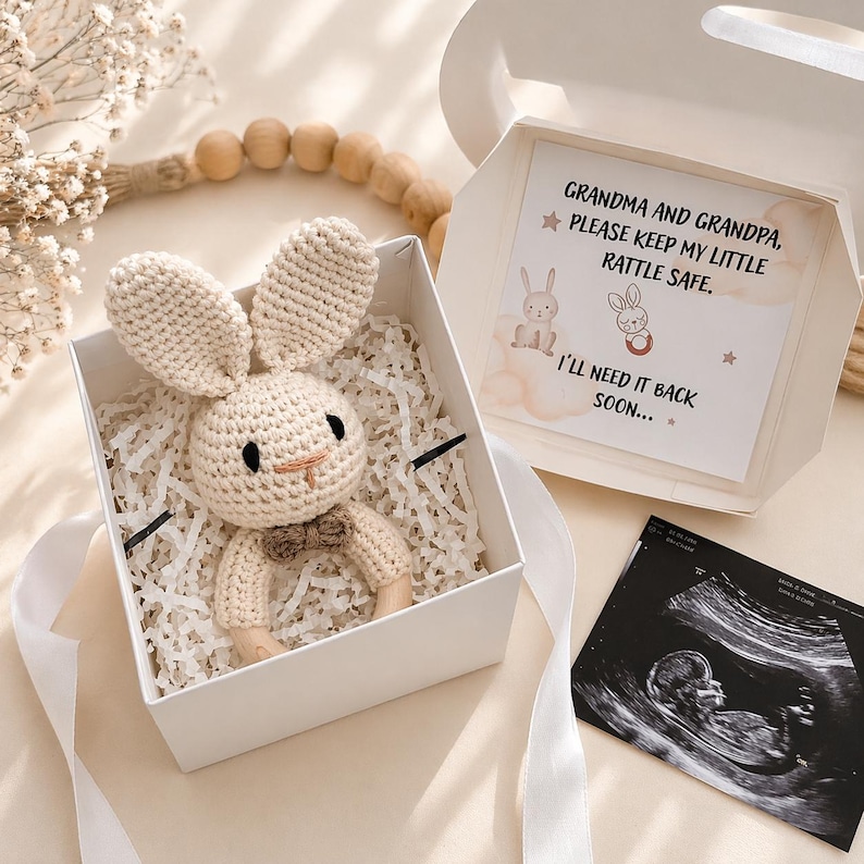 Puede incluir: Un sonajero de conejo de ganchillo beige con una peque&ntilde;a pajarita marr&oacute;n dentro de una caja de regalo blanca. La caja contiene una tarjeta con el texto: GRANDMA AND GRANDPA, PLEASE KEEP MY LITTLE RATTLE SAFE. I'LL NEED IT BACK SOON... y una ecograf&iacute;a.