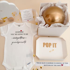 May include: A white onesie with the text "SURPRISE! YOU'RE GOING TO BE babysitters grandparents" sits next to a white box with a gold balloon inside. A white tray with "POP IT" in gold lettering and a safety pin is also present.