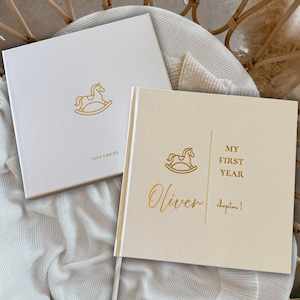 May include: Two baby memory books with gold foil rocking horse illustrations. One book is white with "TINY KICKS" text, the other is cream-colored with "Oliver" and "MY FIRST YEAR" text. Both books are square and sit on a white blanket.