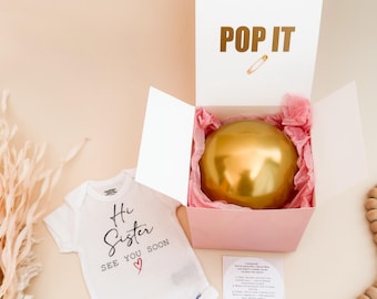 Puede incluir: Un body blanco para bebé con el texto negro Hi Sister SEE YOU SOON y un corazón rojo, junto a una caja de regalo rosa abierta que contiene una esfera dorada en papel de seda rosa. La tapa tiene el texto dorado POP IT y un imperdible.