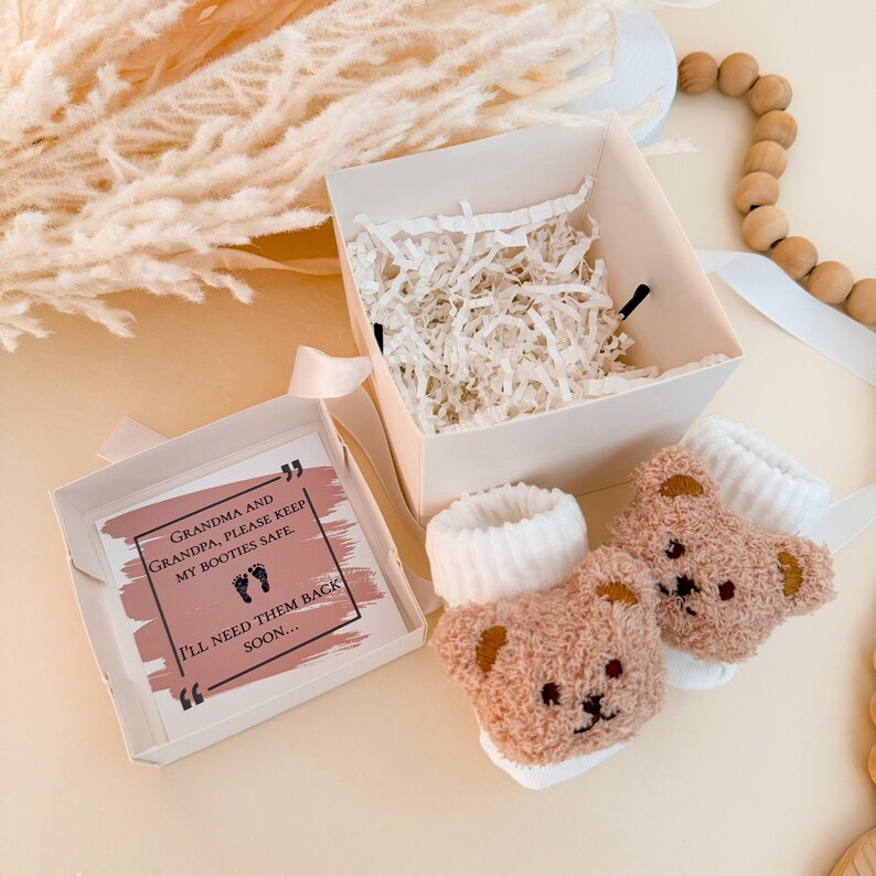 Puede incluir: Un par de patucos para beb&eacute; de peluche en color marr&oacute;n claro con dise&ntilde;o de oso y pu&ntilde;os blancos acanalados, acompa&ntilde;ados de una caja de regalo cuadrada con relleno de papel blanco y una tarjeta con el texto: Grandma and Grandpa, please keep my booties safe. I'll need them back soon.