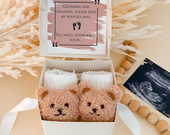 Puede incluir: Una caja de regalo blanca que contiene dos patucos de bebé de peluche color canela con forma de oso y puños blancos acanalados. La tapa incluye una tarjeta con el texto: Grandma and Grandpa, please keep my booties safe. I'll need them back soon.
