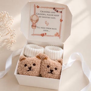 Peut inclure: Une boîte cadeau blanche contenant deux paires de chaussons pour bébé et deux paires de chaussettes blanches. Les chaussons ont la forme d'ours en peluche. Le couvercle de la boîte porte l'inscription "Grand-mère et grand-père, gardez mes chaussons en sécurité. J'en aurai besoin bientôt..."