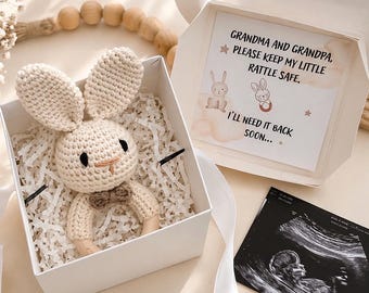 Puede incluir: Un sonajero de conejito de ganchillo en una caja de regalo blanca con papel triturado. El conejito tiene orejas largas, una pajarita y un anillo de madera. Una tarjeta dentro de la caja dice: "Abuela y abuelo, por favor, mantengan mi pequeño sonajero a salvo. Lo necesitaré pronto..." También hay una imagen de ultrasonido.