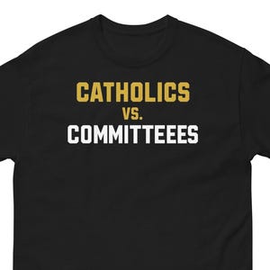 Op de afbeelding: Zwart T-shirt met de tekst "CATHOLICS vs. COMMITTEES" in gouden en witte blokletters. Het T-shirt heeft een ronde hals.