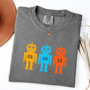 May include: Gray Comfort Colors t-shirt with three colorful robot graphics. The robots are yellow, blue, and orange, each with a number: 01, 10, and 11. A gold necklace with a pendant is draped across the shirt.